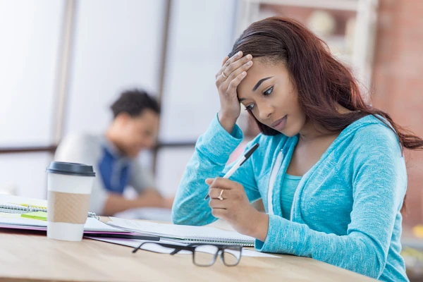 Student sitting at desk doing school work, hand on forehead looking stressed; another student studying (out of focus in background)