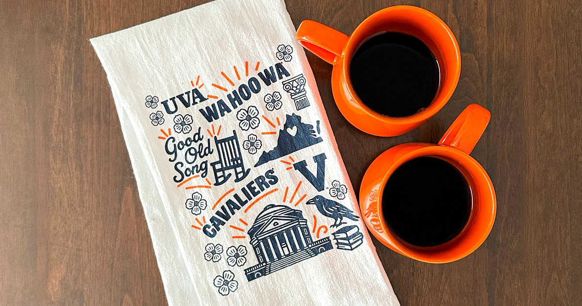 The image shows two orange coffee mugs filled with black coffee next to a white towel featuring decorative University of Virginia-related designs and text.