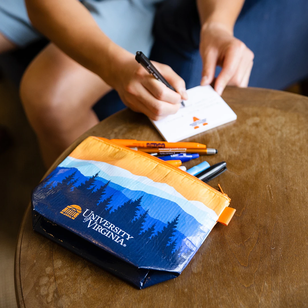 A person is writing on a notepad beside a University of Virginia pencil case and assorted pens.